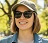 A woman wearing sunglasses and a baseball cap, smiling while standing outside on a sunny day.