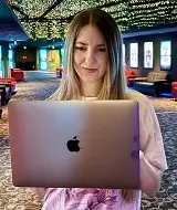 A young woman, identified as the CEO of biteofcode.com, is holding an open MacBook laptop towards the camera with a slight smile. She is wearing a light-colored t-shirt. The background appears to be a dimly lit cinema or theater interior, featuring rows of red seats and ceiling lights that resemble small stars.