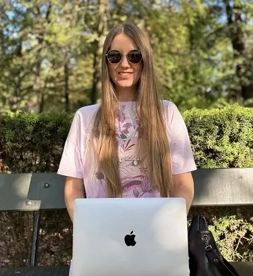This photo shows a woman with long, brown hair sitting on a bench in a park. She is wearing a light pink T-shirt and round-frame sunglasses. An open laptop with the Apple logo is on her lap. The woman is facing the camera and smiling slightly. In the background, there are green trees and bushes, and the sun casts gentle shadows on her.